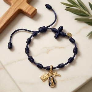 Könnte beinhalten: Ein dunkelblaues Kordelarmband mit einer Reihe von Knoten und drei goldfarbenen Anhängern: einer rechteckigen Plakette, einem kleinen Kreuz und einem ovalen Anhänger, der eine Figur in Blau und Gold darstellt, verziert mit kleinen klaren Steinen.