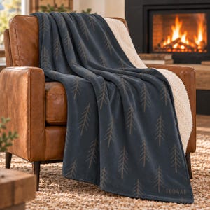 Op de afbeelding: Een donkerblauwe plaid met een herhalend lichtbruin dennenboompatroon en een witte sherpa fleece voering. Het woord SKOGAR staat gedrukt in de onderste hoek van de deken, die over een bruine leren fauteuil is gedrapeerd.