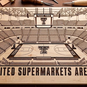 Könnte beinhalten: Eine rechteckige, helle Holzplatte mit einer detaillierten, lasergravierten Illustration des Basketballfeldes der United Supermarkets Arena. Das Design zeigt das Texas Tech-Logo auf der Anzeigetafel und dem Spielfeld sowie den eingravierten Text UNITED SUPERMARKETS ARENA am unteren Rand.