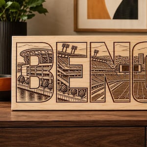 May include: A rectangular light-colored wooden sign featuring laser-engraved illustrations of a football stadium integrated into the large block letters B, E, N, and G. The detailed artwork depicts stadium seating, field lines, and light towers.