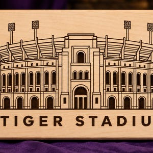May include: A rectangular light-colored wooden plaque featuring a dark brown laser-engraved line drawing of a large stadium. The text TIGER STADIUM is printed in a bold, dark brown sans-serif font centered below the stadium illustration.