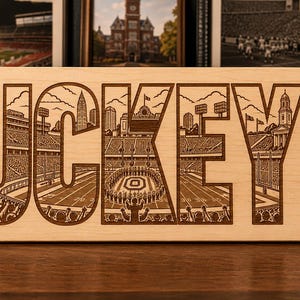 Puede incluir: Un letrero rectangular de madera clara con la palabra BUCKEYES grabada con láser en letras grandes. Cada letra contiene ilustraciones detalladas de un estadio de fútbol, un horizonte urbano y hojas de castaño de Indias.