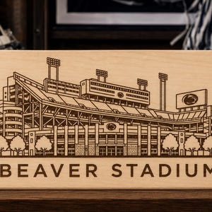 Könnte beinhalten: Eine rechteckige, helle Holzplatte mit einer dunkelbraunen, lasergravierten Abbildung des Beaver Stadiums. Das detaillierte architektonische Design umfasst Zuschauerränge, Flutlichtmasten und den Schriftzug BEAVER STADIUM am unteren Rand.