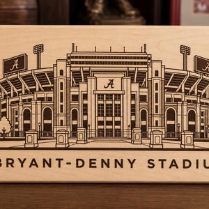 May include: A rectangular light-colored wooden plaque featuring a dark brown laser-engraved illustration of Bryant-Denny Stadium. The design includes the stadium architecture, the letter A on the scoreboards, and the text BRYANT-DENNY STADIUM.