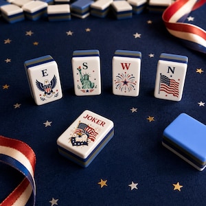 Puede incluir: Un conjunto de fichas blancas y azules con dise&ntilde;os patri&oacute;ticos, incluyendo un &aacute;guila, la Estatua de la Libertad, fuegos artificiales y la bandera estadounidense. Una ficha dice "JOKER". Las fichas est&aacute;n sobre una superficie azul oscuro con confeti en forma de estrella y cinta tricolor.