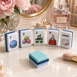 Op de afbeelding: Een set witte en blauwe Mahjong-tegels met kleurrijke illustraties van een macaron, koffiebekers, een handtas en een cadeaukaart. De tegels zijn gerangschikt op een reflecterend oppervlak, met een blauwe tegel op de voorgrond. De tegels zijn genummerd 1, 3, 9 en "JOKER".