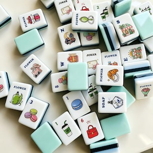 Op de afbeelding: Een verzameling witte en lichtblauwe Mahjong-tegels met diverse illustraties, waaronder bloemen, eten en accessoires. Sommige tegels hebben cijfers en letters. Het woord "FLOWER" staat op meerdere tegels.
