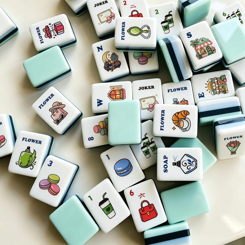 Op de afbeelding: Een verzameling witte en lichtblauwe Mahjong-tegels met diverse illustraties, waaronder bloemen, eten en accessoires. Sommige tegels hebben cijfers en letters. Het woord "FLOWER" staat op meerdere tegels.