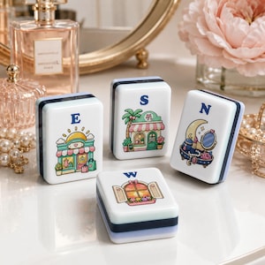 Op de afbeelding: Vier witte en blauwe decoratieve blokken met illustraties en de letters E, S, N en W. De illustraties tonen een winkel, een palmboom, een maan met een spiegel en een raam met bloemen. De blokken zijn rechthoekig.