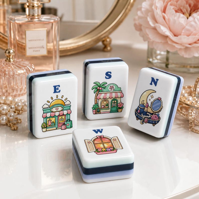 Op de afbeelding: Vier witte en blauwe decoratieve blokken met illustraties en de letters E, S, N en W. De illustraties tonen een winkel, een palmboom, een maan met een spiegel en een raam met bloemen. De blokken zijn rechthoekig.