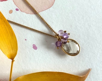 May include: A gold chain necklace with a clear oval pendant and small purple gemstones. The necklace is on a white background with yellow leaves.