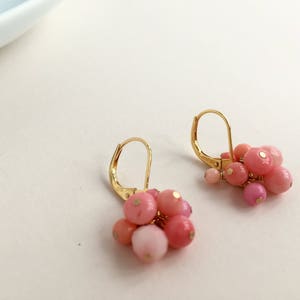 Puede incluir: Un par de pendientes de oro con cuentas rosas y blancas. Los pendientes son de estilo colgante y tienen un racimo de cuentas en la parte inferior.