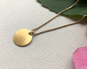 Può includere: Una collana color oro con un piccolo pendente rotondo. Il pendente ha una finitura in metallo spazzolato. La catena è un delicato stile a catena a scatola. Lo sfondo include una foglia verde e un petalo di fiore rosa.