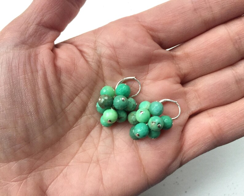 Puede incluir: Un par de pendientes de aro de plata con cuentas de piedras preciosas verdes. Las cuentas est&aacute;n agrupadas en forma de flor.