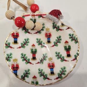 May include: A decorative scallop shell ornament featuring a repeating pattern of nutcrackers and pine branches on a white background. The shell has a gold-painted edge and is topped with a red and white Santa hat charm and wooden beads.