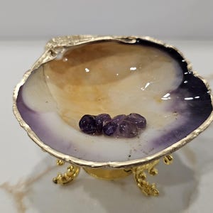May include: A decorative seashell dish with a polished interior featuring shades of cream, tan, and deep purple. The shell edge is trimmed in gold, and it rests on a small, ornate gold-toned metal stand. Inside the shell are several small, tumbled purple amethyst stones.