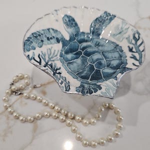 May include: A decorative scallop shell dish featuring a hand-painted blue sea turtle and coral motif on a white background with a silver-toned rim, accompanied by a strand of white pearls arranged in a circular pattern below the shell dish.