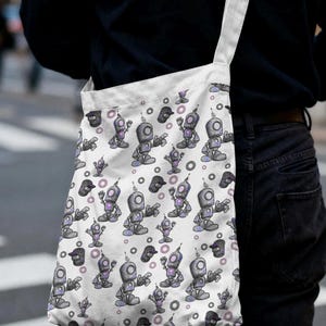 Puede incluir: Un bolso de tela blanco con un patrón repetitivo de pequeños robots grises con detalles brillantes en color púrpura y formas circulares. El bolso cuenta con una correa larga para el hombro y está hecho de tela resistente con fondo claro.
