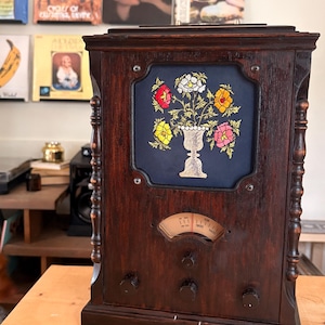 May include: A vintage wooden radio with a dark wood finish. The radio has a navy blue panel with an embroidered floral design in a vase. The radio has knobs and a dial, and is set on a wooden table. The background includes album covers.