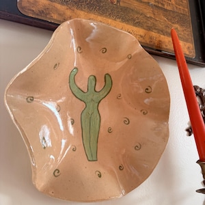 May include: A beige ceramic bowl with a wavy, irregular rim. The center features a green stylized figure with raised arms, surrounded by small green spiral patterns scattered across the interior surface of the bowl.