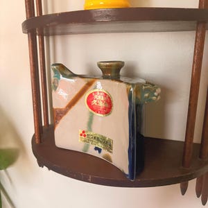 May include: A square ceramic container with a lid, featuring a beige, blue, and brown color scheme. The container has a small handle on the side and a round, brown knob on top. It sits on a dark brown wooden shelf, with a yellow pitcher above.