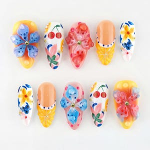 May include: A set of ten press-on nails featuring almond-shaped tips with varied designs including 3D blue and pink floral accents, hand-painted cherries, citrus slices, and yellow sunburst patterns, all embellished with small gold metallic studs.