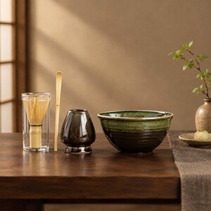 May include: A matcha tea set including a bamboo whisk inside a clear glass cylinder, a bamboo scoop, a dark brown ceramic whisk holder, and a wide, deep green and black glazed ceramic bowl with a textured, ribbed exterior surface.