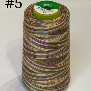 May include: A large spool of variegated sewing thread featuring a multi-coloured pattern of yellow, green, blue, and red stripes. The top of the spool has a green plastic cap with the printed text #5 and 40/2. The thread is wound in a conical shape.