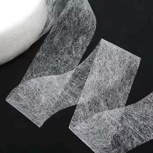 May include: A roll of white, semi-transparent fusible interfacing tape with a fibrous, non-woven texture. The tape is shown partially unrolled, displaying its lightweight, mesh-like construction and consistent width across the entire length.