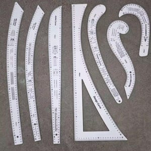 May include: A collection of white plastic dressmaking rulers, including curved and straight edges, and a set square. Each ruler has markings in both centimetres and inches, with diagrams of clothing patterns.