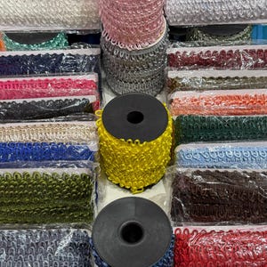 May include: A collection of assorted loop fringe trim spools in various colours including yellow, pink, grey, blue, red, green, and brown. Each spool features a decorative braided edge design, with several rolls individually wrapped in clear plastic.