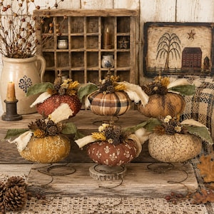 May include: Six fabric pumpkins in various patterns and colors, each topped with pinecones, dried wheat, and frayed fabric leaves. The pumpkins are mounted on metal springs and wooden bases, arranged on a rustic wooden surface with a wooden grid.