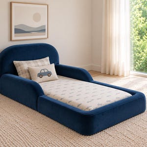 May include: A navy blue velvet toddler bed with a rounded headboard and integrated side rails. The bed features a white mattress with a repeating blue car pattern, a beige plaid pillow, and a small rectangular pillow with a blue car graphic.