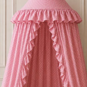 May include: A pink fabric canopy featuring a white polka dot pattern, a ruffled trim along the top edge, and a pointed conical top. The canopy hangs from a single point and drapes down to cover a round, tufted cream-colored floor cushion.