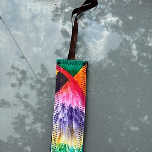 May include: A rectangular fabric bookmark featuring a vibrant, multi-colored hand-drawn design with shades of green, orange, pink, purple, and yellow. The edges are finished with black zigzag stitching, and a brown ribbon loop is attached to the top.