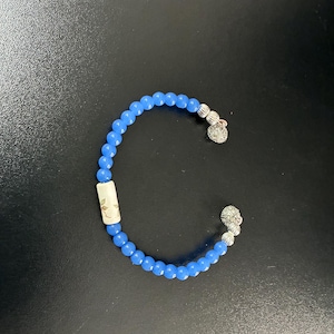 May include: A blue beaded bracelet featuring a series of small, round, translucent blue beads. The bracelet includes a single cylindrical off-white bead with a delicate brown leaf pattern and two silver-toned textured end caps with charms.