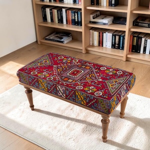 May include: A rectangular ottoman featuring a vibrant red fabric upholstery with a multi-coloured geometric pattern. The piece is supported by four turned wooden legs in a light brown finish, providing a stable base for the cushioned seat.