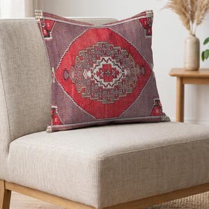Puede incluir: Un cojín cuadrado que presenta un medallón central en forma de diamante rojo con intrincados patrones geométricos en tonos gris, blanco y negro. El fondo del cojín es de un color malva-púrpura apagado con acentos geométricos a juego en las esquinas.