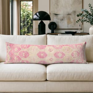 May include: A long rectangular lumbar pillow featuring a faded pink and cream geometric pattern. The textile displays a repeating hexagonal motif with intricate tribal-inspired details across the entire surface of the elongated cushion cover.
