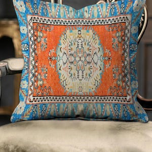 Könnte beinhalten: Ein quadratisches Zierkissen mit einer leuchtend orangefarbenen Mittelplatte, einem hellcremefarbenen Medaillon und komplexen geometrischen Mustern in Blau, Schwarz und Weiß. Der Rand zeigt ein wiederkehrendes blau-weißes Motiv mit Verzierungen.