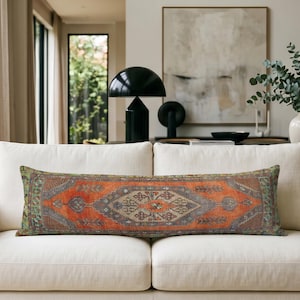 Puede incluir: Un cojín lumbar rectangular largo que presenta un patrón tejido tradicional en tonos naranja quemado, gris pizarra y verde apagado. El textil muestra un diseño de medallón central con bordes geométricos intrincados.