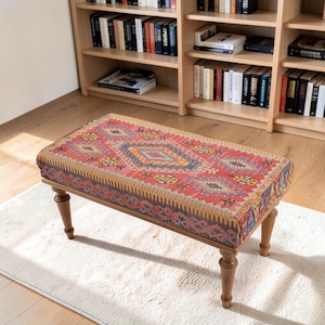 May include: A rectangular ottoman featuring a vibrant red, orange, and blue geometric patterned fabric upholstery. The piece is supported by four turned wooden legs with a medium brown finish, providing a stable base for the cushioned seat top.