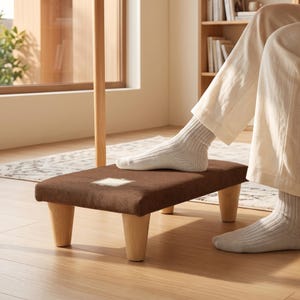 May include: A rectangular brown fabric-covered footstool with four light-coloured wooden legs. The top surface features a small, centred square patch with a grid pattern. The footstool is positioned on a wooden floor, supporting a pair of feet.