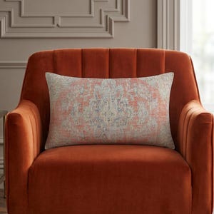 May include: A rectangular throw pillow featuring a faded, intricate medallion pattern in shades of burnt orange, muted blue, and cream. The pillow has a textured, woven appearance and is centered on a solid burnt orange velvet armchair.