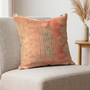 Puede incluir: Un cojín cuadrado con un patrón descolorido de inspiración vintage en tonos coral, beige y verde salvia suave. El diseño incluye un panel central vertical con motivos geométricos, rodeado por un borde y formas angulares.