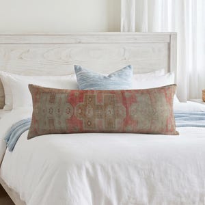 Könnte beinhalten: Ein langes, rechteckiges Lendenkissen mit einem verblassten geometrischen Muster in Rot und Beige. Das Textil zeigt ein sich wiederholendes Motiv mit gedämpften salbeigrünen Akzenten und einem verwitterten Aussehen, mittig auf dem Bett.