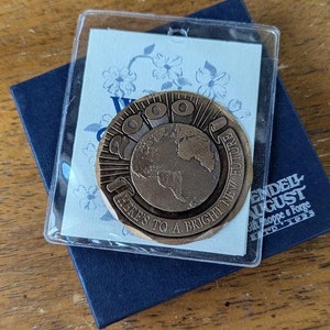 May include: A bronze-colored commemorative coin with the text "2010" and an image of the Earth. The coin is in a clear plastic case, resting on a blue card with floral designs and a navy blue box with the text "Wendell August Gift Shoppe & Forge ESTD 1923".
