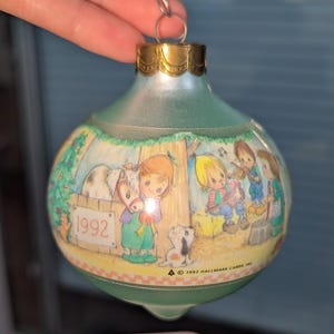 Könnte beinhalten: Ein Vintage-Weihnachtsschmuck mit einem grünen Glaskörper und einer goldfarbenen Metallkappe. Der Schmuck zeigt eine gemalte Szene von Kindern mit einem Pferd, einem Hund und Musikern. Das Jahr "1992" ist auf dem Ornament sichtbar.