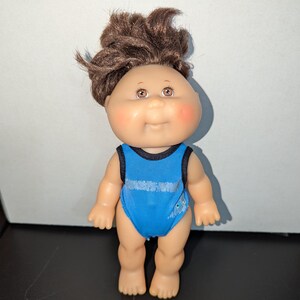May include: A small Cabbage Patch Kid doll with brown hair wearing a blue swimsuit with a black trim.