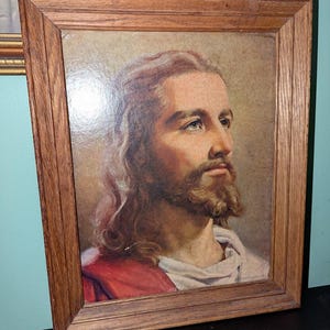 May include: A framed portrait of a man with long brown hair and a beard, wearing a white robe and a red garment. The portrait is in a wooden frame.